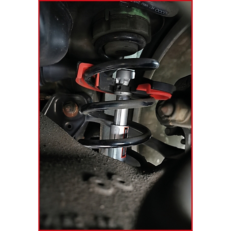 Nahaufnahme Stoßdämpfer mit roter Klemme. Schwarze Feder und Metallhülse mit rotem Logo. Schrauben und weitere Fahrzeugteile im Hintergrund.